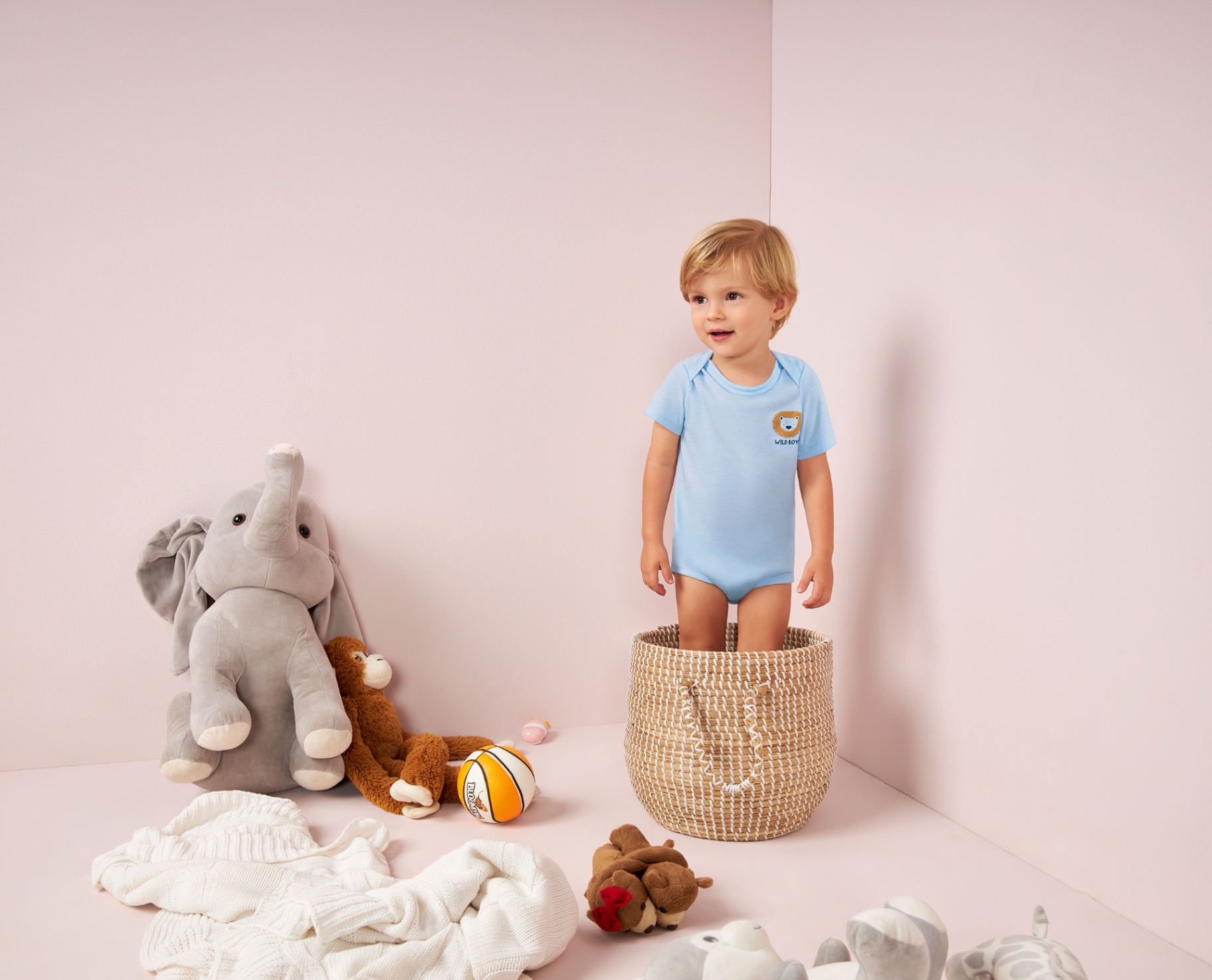 Toddler in blue onesie standing in basket among plush toys